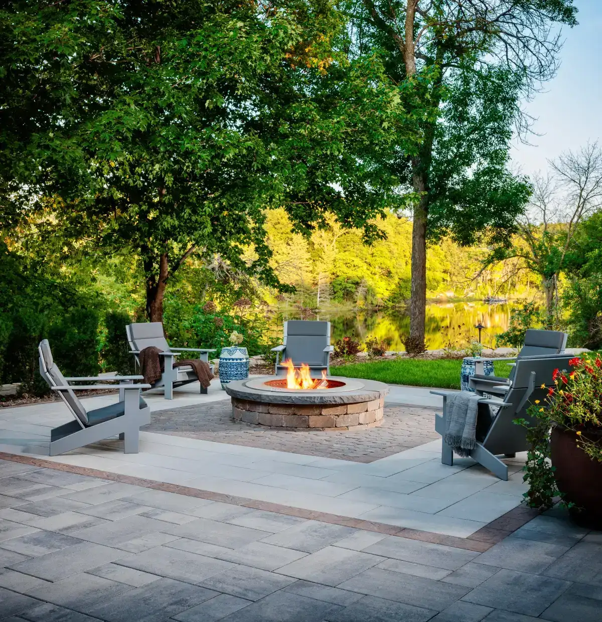 Round fire pit with four chairs on a paver patio beside trees and water